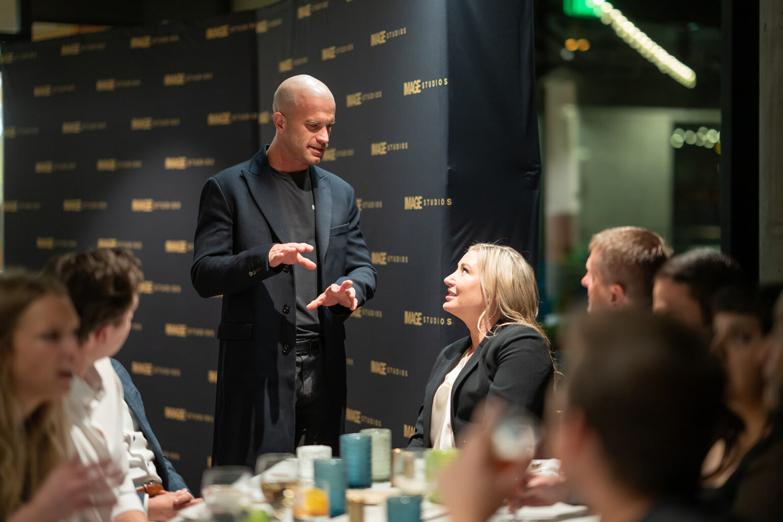 A man in a black coat is speaking at a table surrounded by people