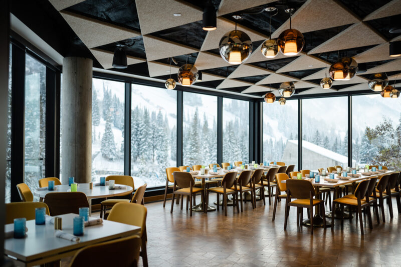 A modern restaurant dining room featuring large windows with a mountain view. Tables are set, and unique lighting fixtures hang from a geometric ceiling