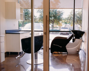 A salon room with a shampoo bowl and swivel chair is displayed. The room has a white counter and view of the outdoors