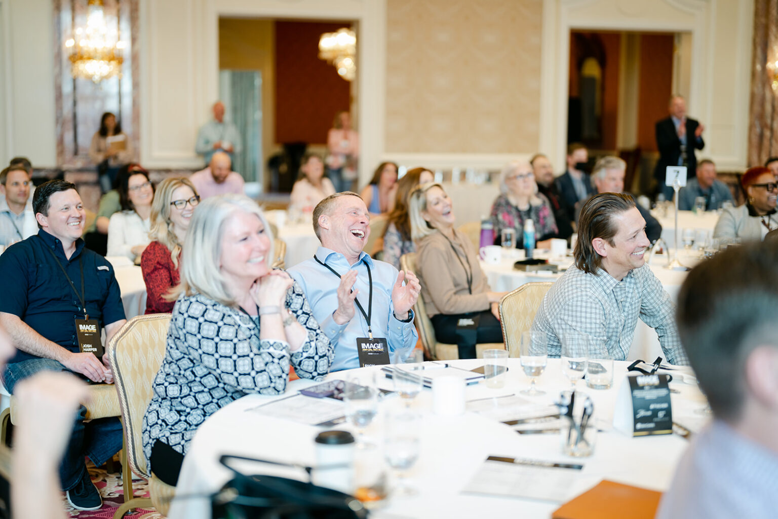 A professional audience sits at round tables, laughing and clapping during a presentation